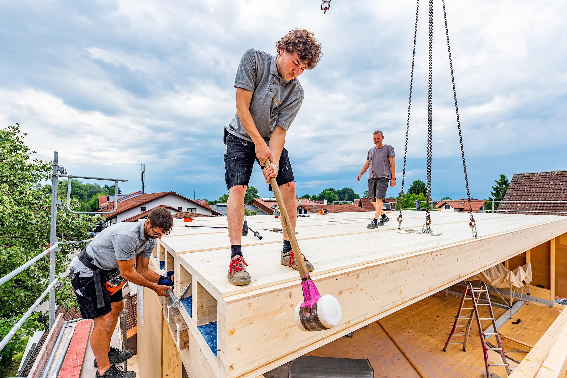Montage sur chantier : trois charpentiers assemblent et décrochent les éléments CLT BOX posés en toiture, élingues de grue encore en place