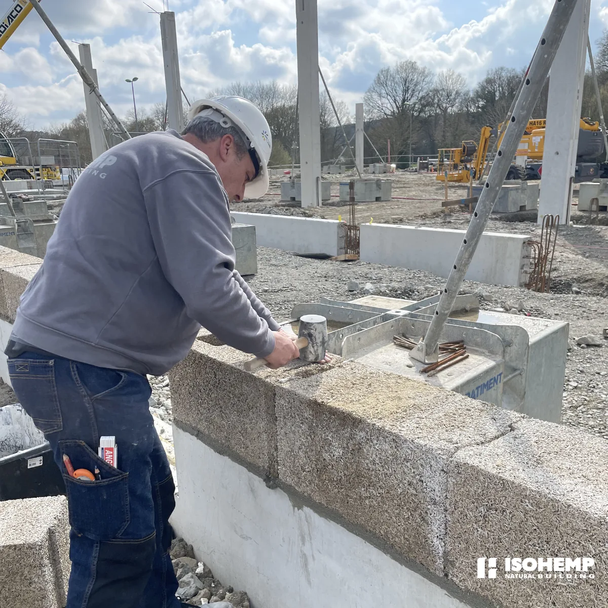 Macon casque appliquant le mortier-colle a la truelle sur une arase de blocs de chanvre, grue et chantier du magasin LIDL de La Gacilly en arriere-plan