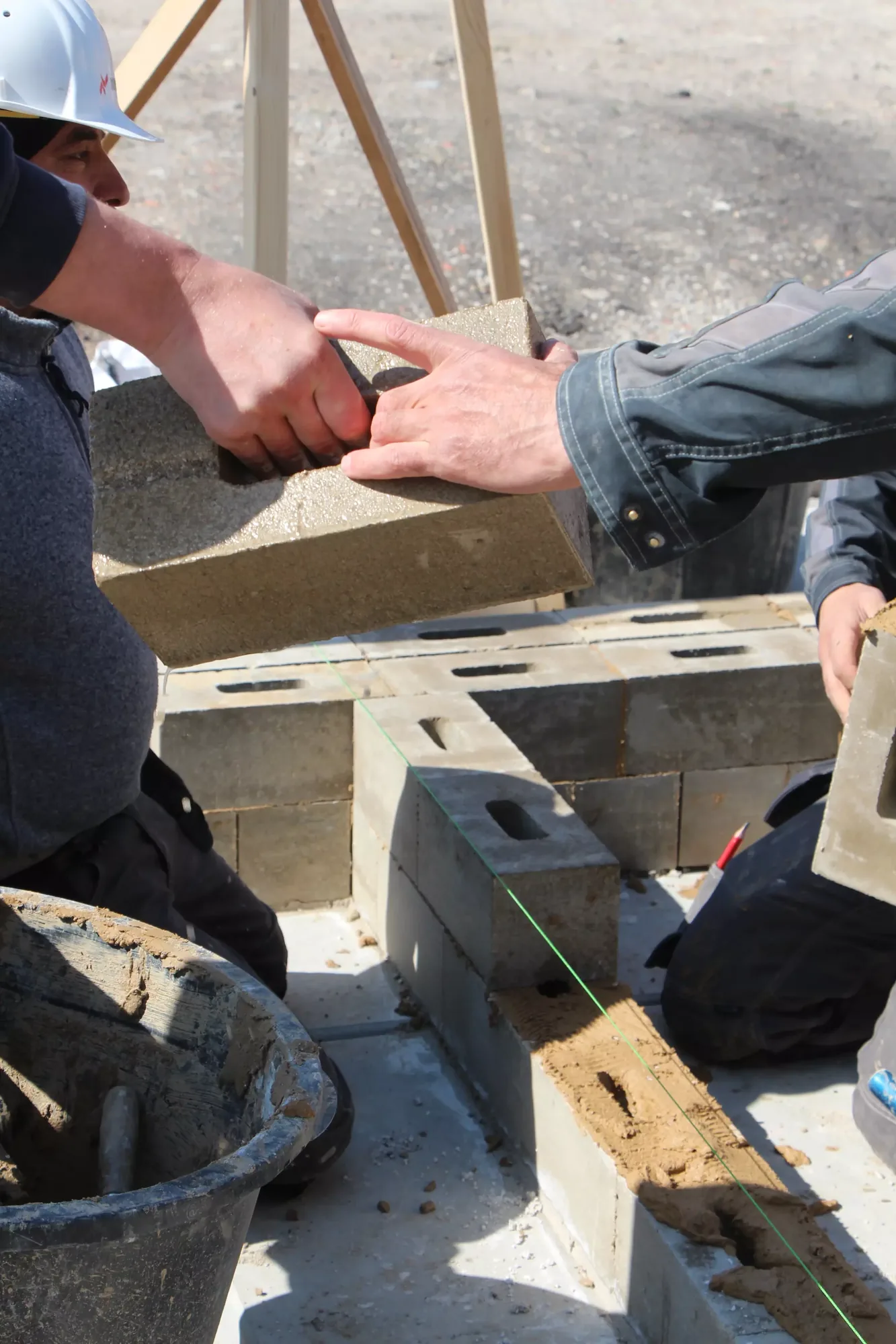 Maçons casqués posant des blocs de terre comprimée sur chantier, mains en gros plan, seau de mortier de terre au premier plan