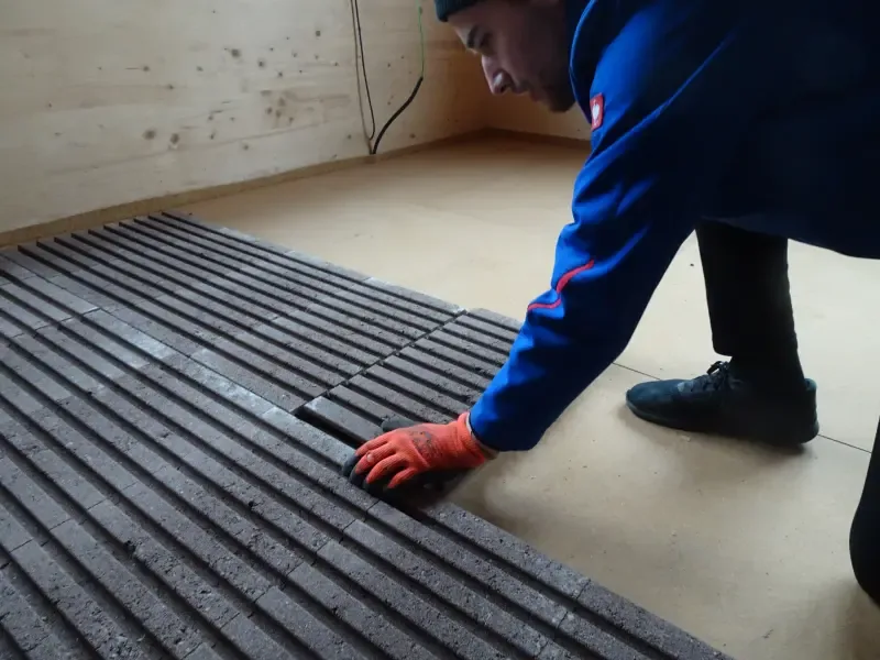 Ouvrier gante posant une dalle Lithotherm a la main sur le support, dans une piece a ossature bois — alt d'origine : 'Ein Handwerker verlegt gerillte Bodenplatten auf einer Holzunterkonstruktion'