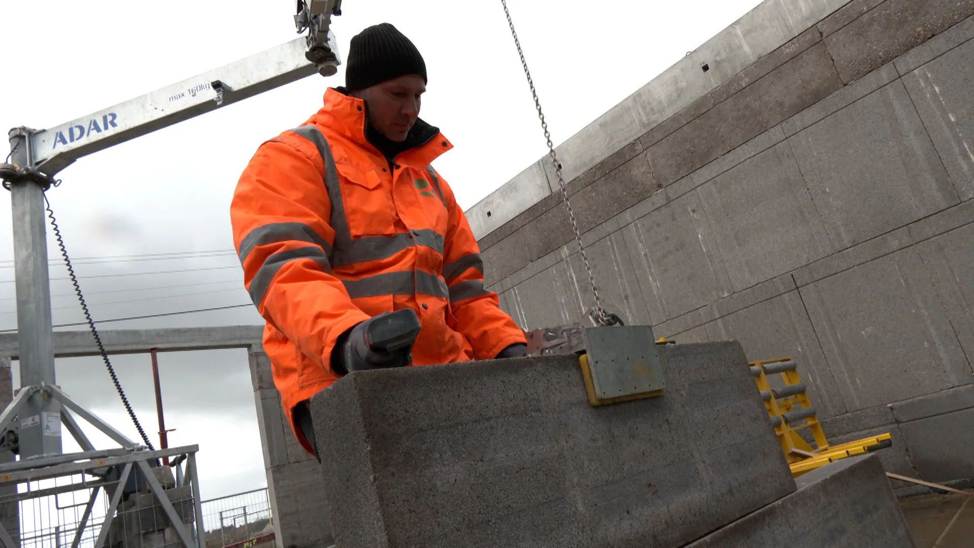 Chantier-test Roosens Bétons : pose d'un élément de mur préassemblé à la mini-grue, mur en éléments éco-circulaires en arrière-plan (reportage Antenne Centre sur le bloc Clayton)