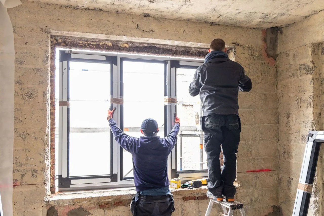 Two installers packing a PVC window frame into a bare masonry opening in a renovation