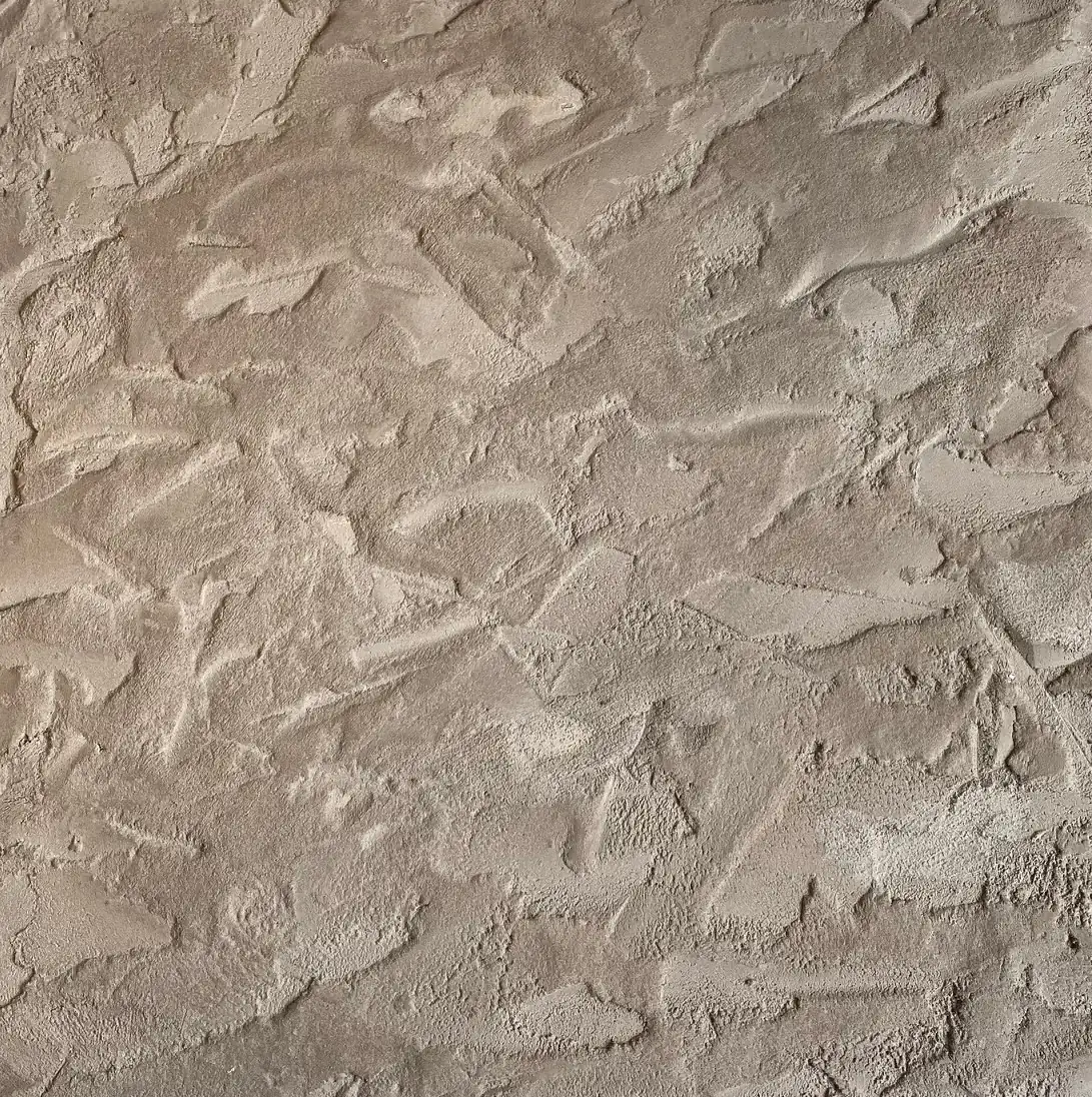 Gros plan sur une finition Claystone effet pierre naturelle (chantier Matonge, Bruxelles) : reliefs de lisseuse, plages lissées et arrachements, teinte sable/taupe en lumière rasante.
