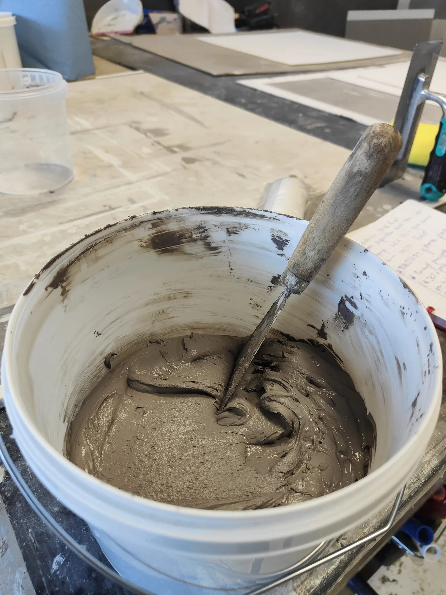 Mise en oeuvre du Claystone lors d'une formation ClayLime : le mortier d'argile fraîchement gaché dans le seau, lisseuse inox plantée dedans, plaques d'essai en cours de séchage a l'arrière-plan.