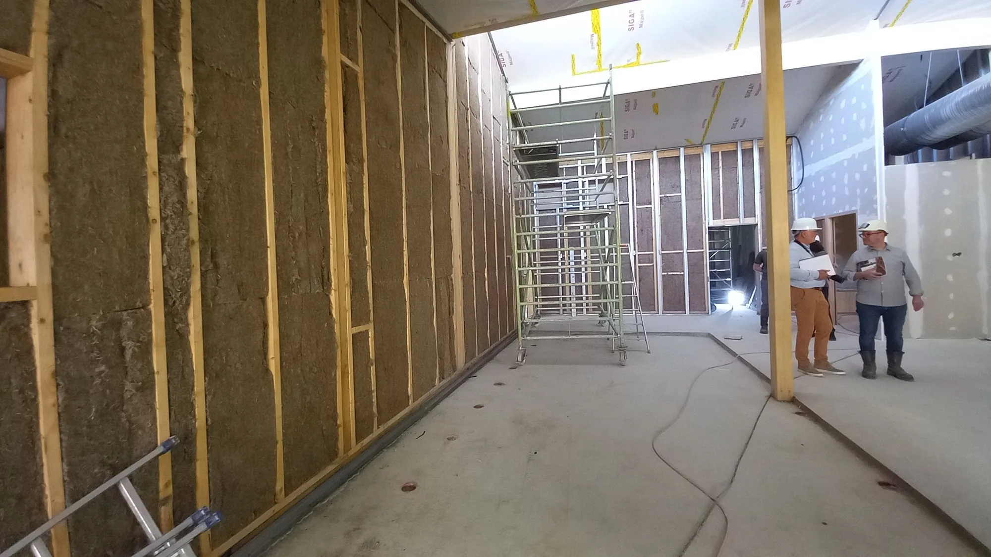 Chantier : mur en ossature bois isole en fibres d'herbe, vue interieure large avec ouvriers au fond