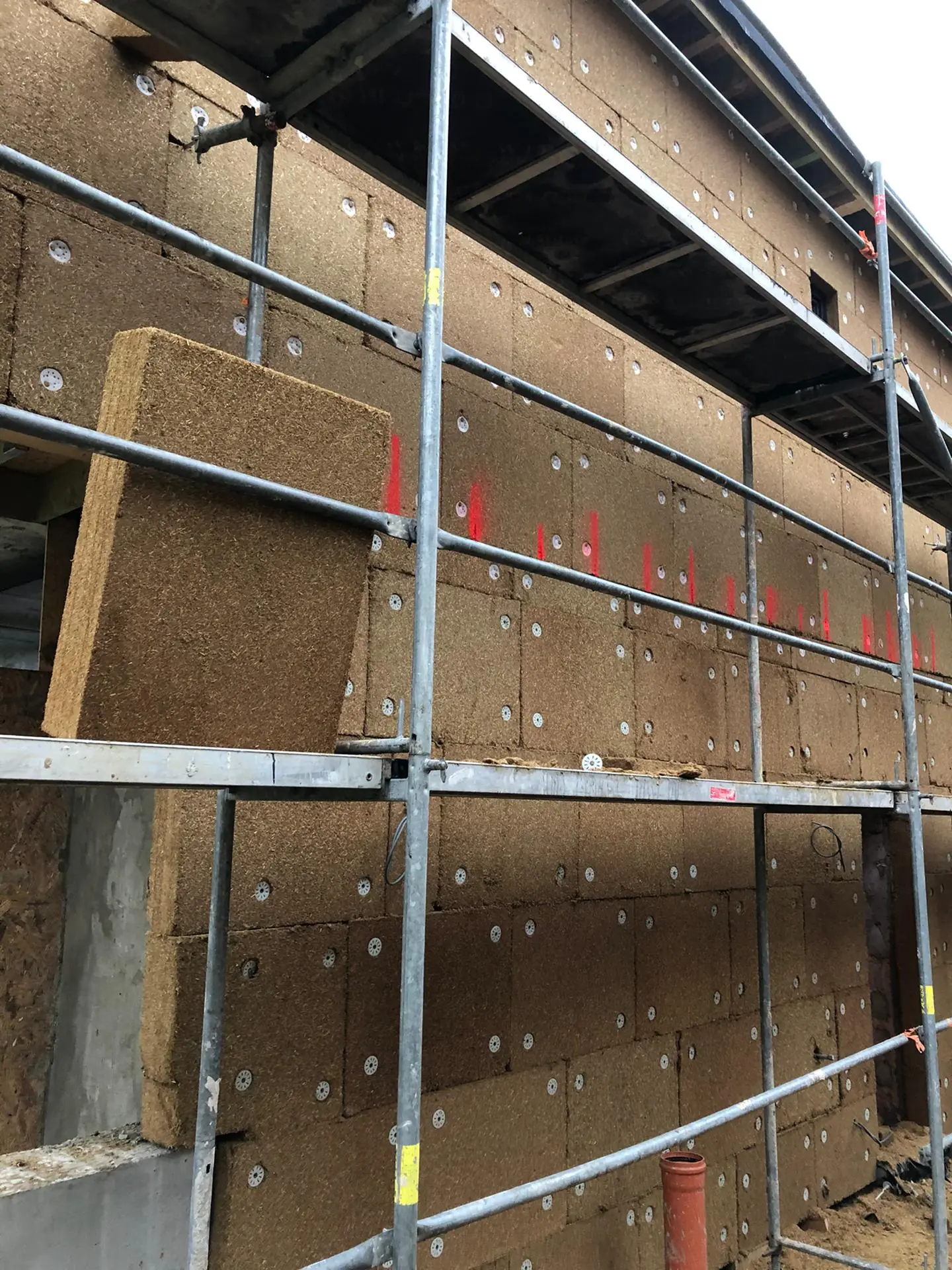 Mise en œuvre en ITE : panneau de paille en attente sur l'échafaudage devant la façade en cours de calage-chevillage (repères rouges, rosaces)