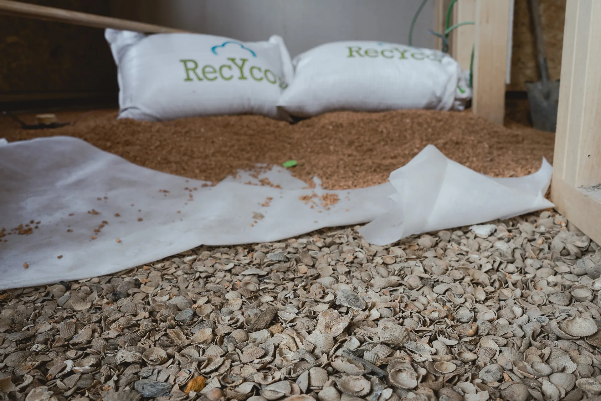 Complexe de sol en détail : granulés Recycork épandus en remplissage, sacs Recycork au fond, lit de coquilles et voile en premier plan