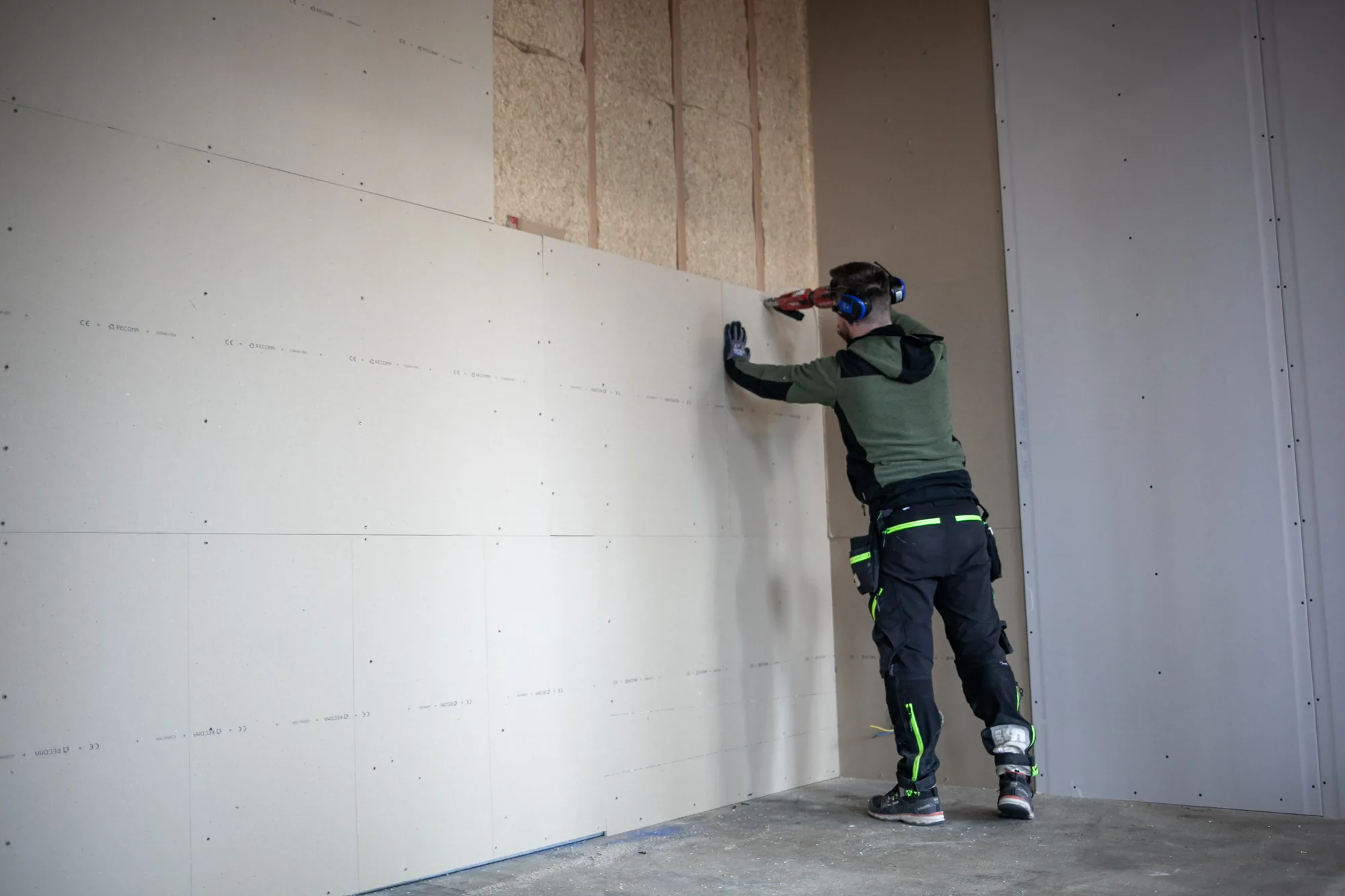 Chantier AMF Fastigheter / SMK Entreprenad (Stockholm, 2024) : vissage des panneaux PackWall sur ossature Wood Tube et isolant chanvre. Le marquage « CE · RECOMA » imprimé sur la face du panneau identifie le matériau, et l'isolant apparaît dans la travée encore ouverte. © Recoma AB