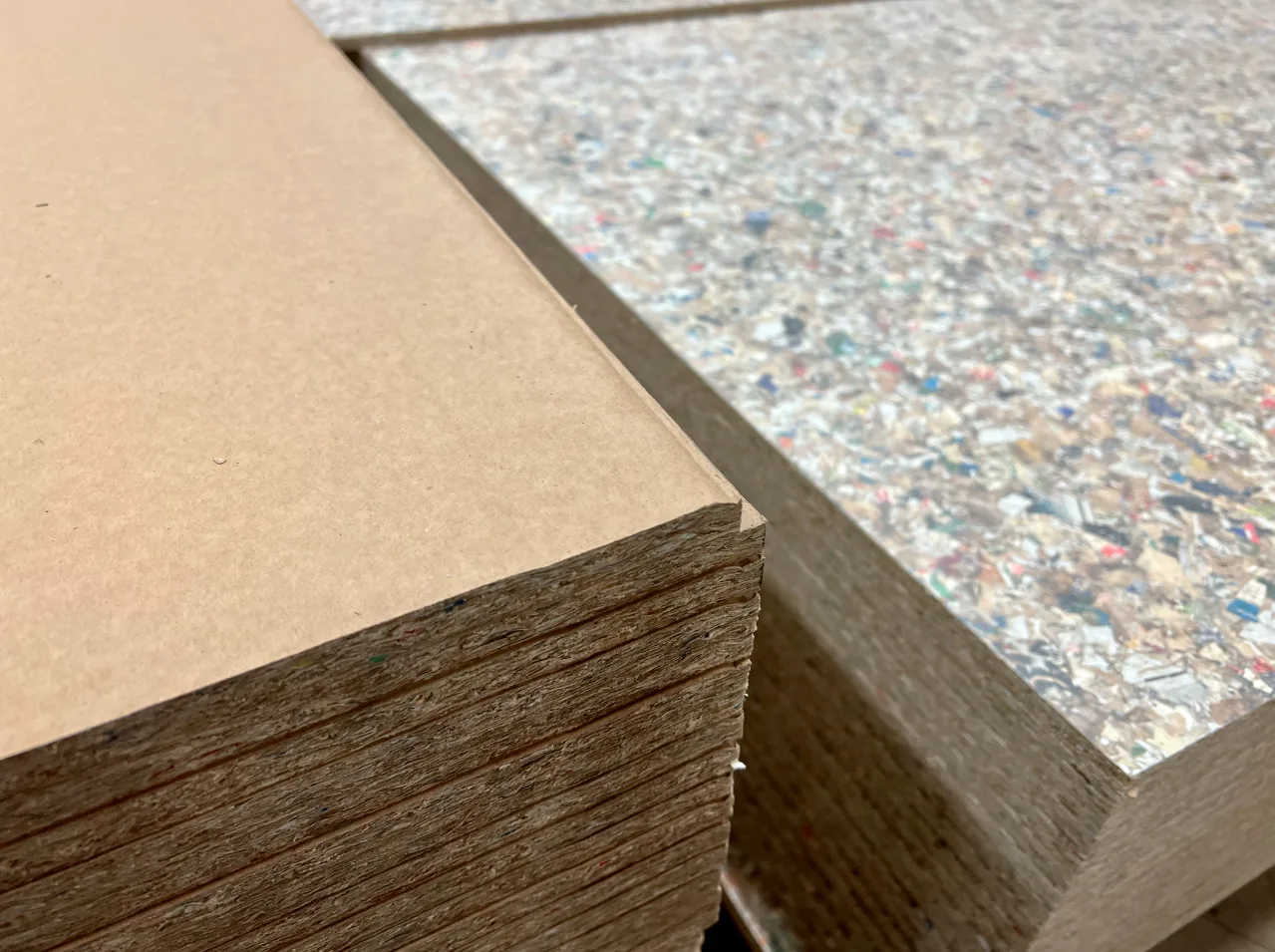 Gros plan sur des panneaux Recoma empilés : à gauche la face lisse kraft (PackWall Basic), à droite la face mouchetée (PackWall Design), et au centre la tranche qui révèle l'épaisseur, le feuilletage des couches compressées et les éclats colorés. © Recoma AB